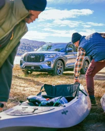 Two people preparing a kayak by a parked blue SUV near a lakeside under a partly cloudy blue sky.