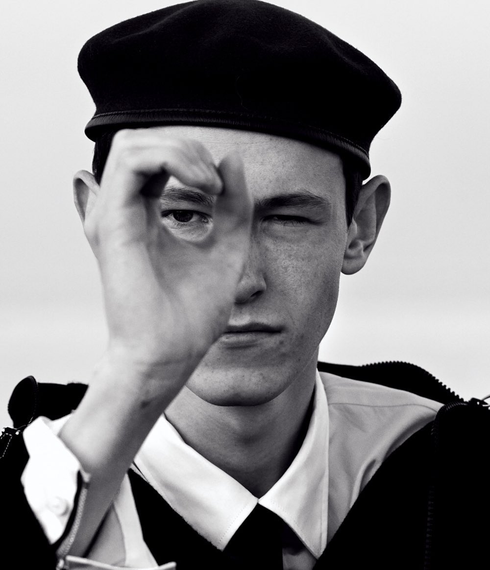 Monochrome portrait of a young adult man wearing a beret, making an 'OK' hand gesture over one eye, winking at the camera in black and white.