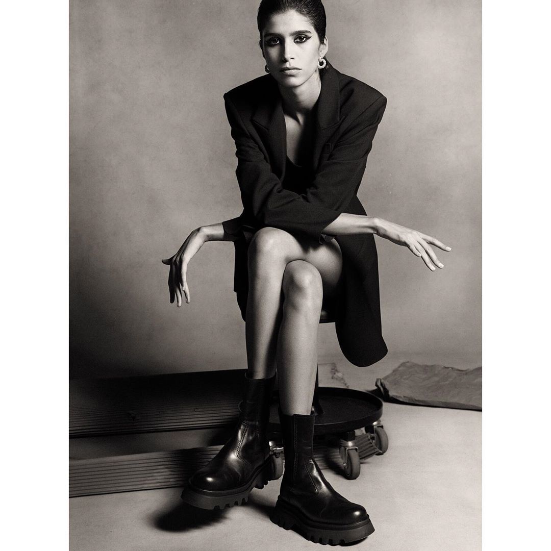 Monochromatic fashion portrait of a woman with dark hair and black boots and blazer sitting on a stool in a photo studio.
