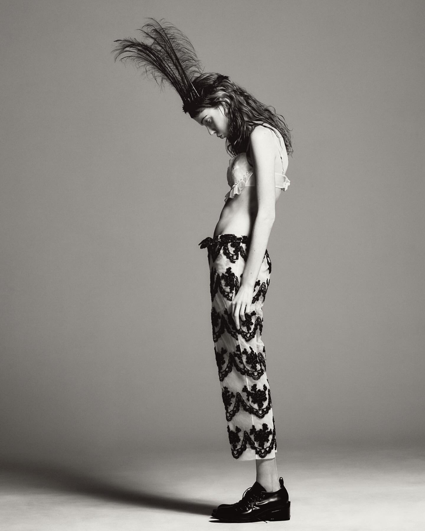 Monochrome portrait of a slender woman in cropped lace pants, bralette, headdress with feathers and platform shoes, looking down in a studio.