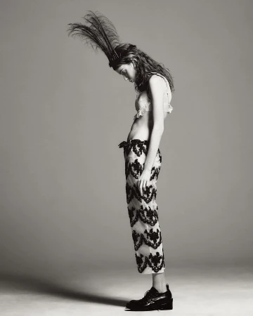Monochrome portrait of a slender woman in cropped lace pants, bralette, headdress with feathers and platform shoes, looking down in a studio.