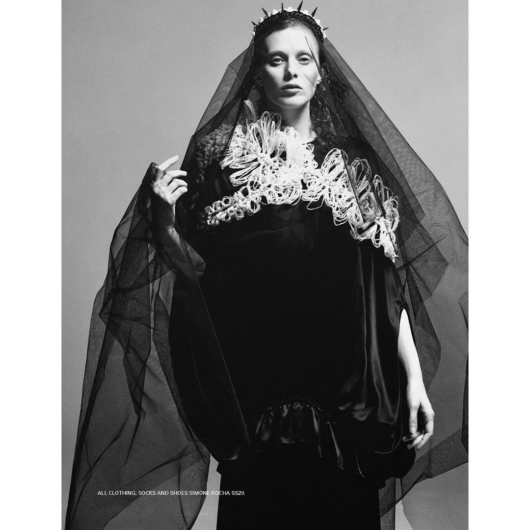 Black and white fashion portrait of a woman wearing an ornate headpiece and veil with a dramatic cape and a dark background.