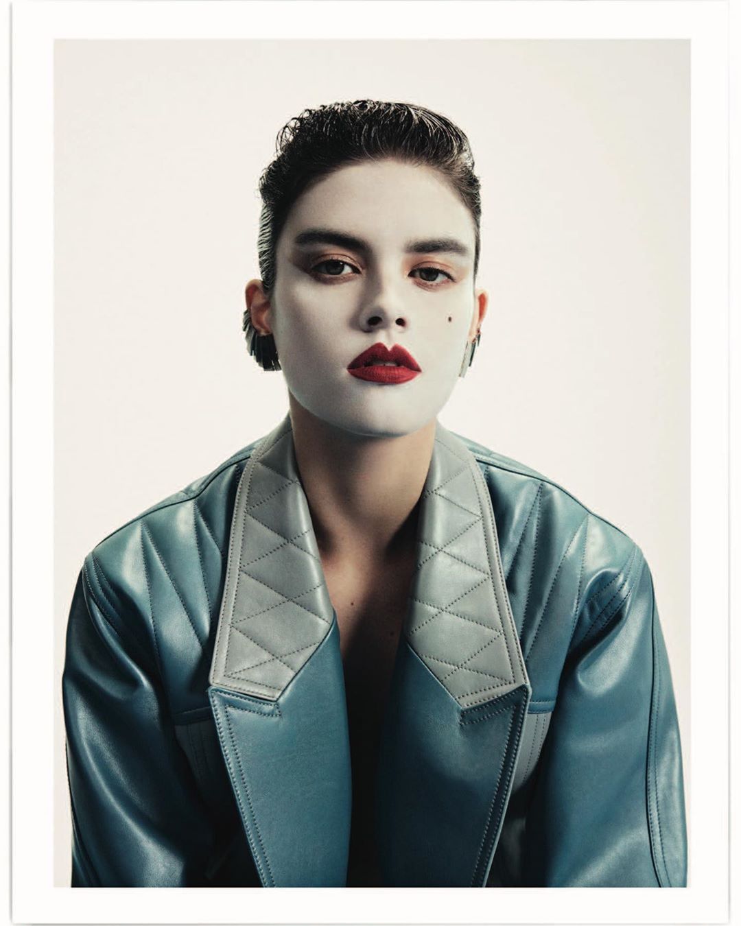 Close-up portrait of an adult woman wearing a blue leather jacket with white makeup and dark red lipstick, posing against a white backdrop.