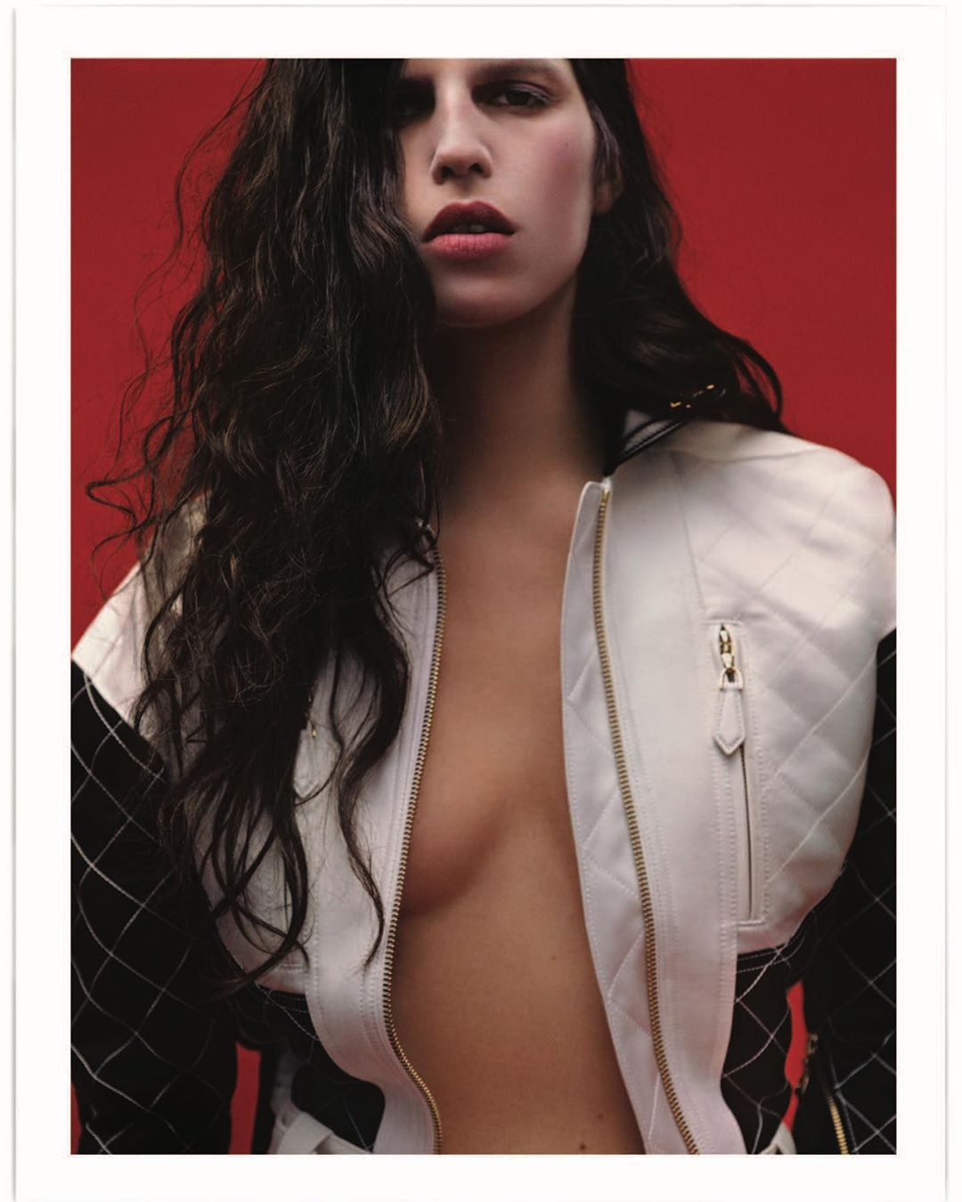 Portrait of a young adult woman with long dark hair wearing a partially unzipped white jacket against a red background.