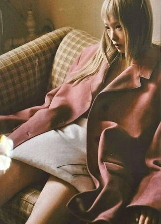 A young adult woman poses on a plaid couch in a pink blazer. Fashion editorial photography with muted tones and soft indoor lighting.