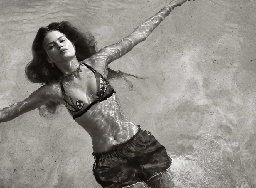 Monochrome shot of a woman in bikini and shorts floating in water, arms outstretched, creating a serene and peaceful atmosphere.