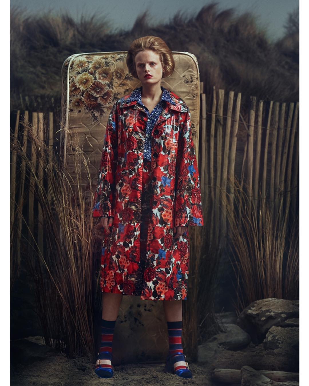 A woman in a floral coat stands outdoors against a weathered mattress, wearing striped socks and dress shoes, with dry grass around.