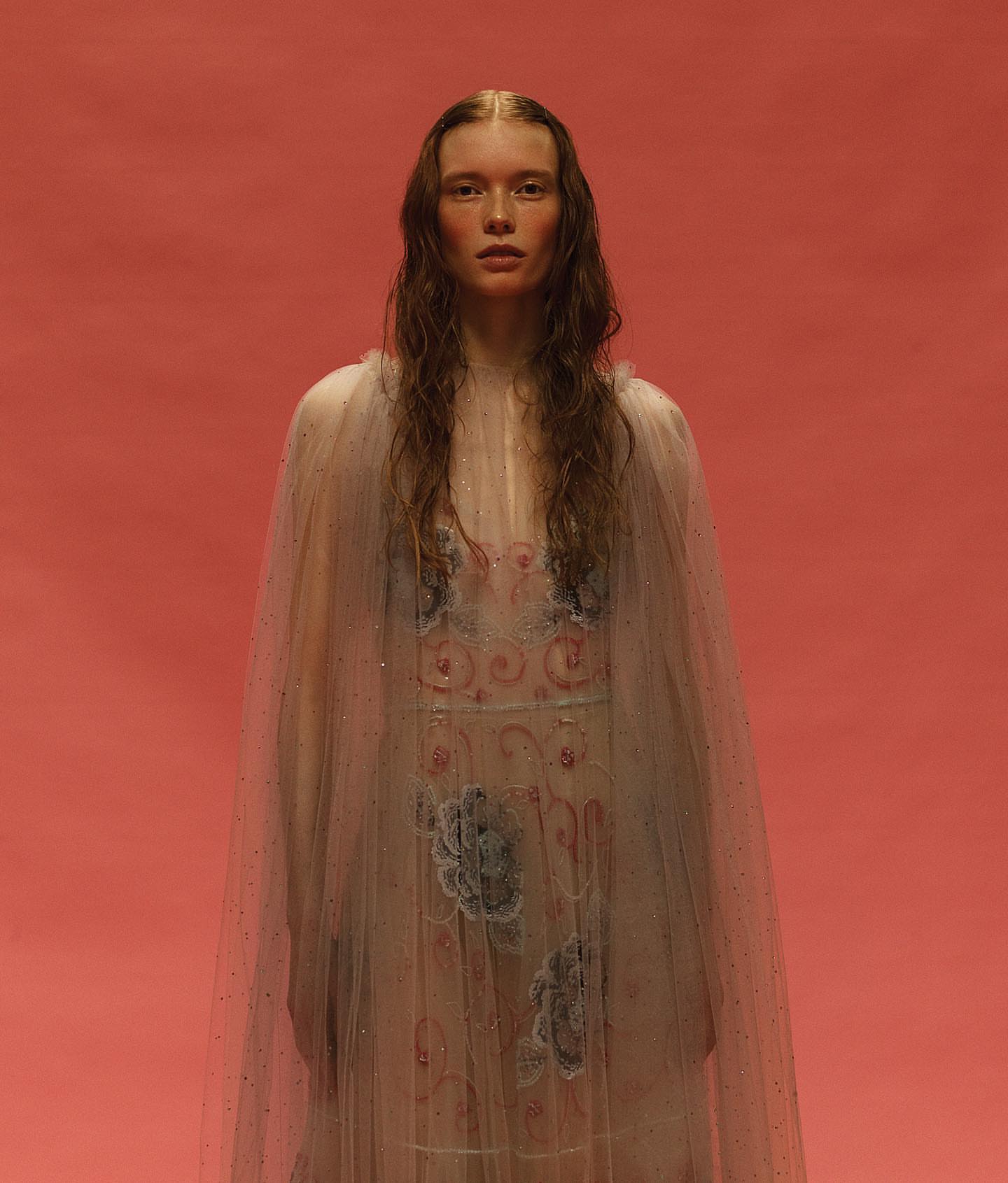 Portrait of a young fair-skinned woman with long brown hair wearing a sheer, decorated cape against a solid salmon-colored background.