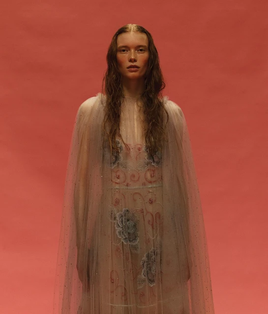 Portrait of a young fair-skinned woman with long brown hair wearing a sheer, decorated cape against a solid salmon-colored background.