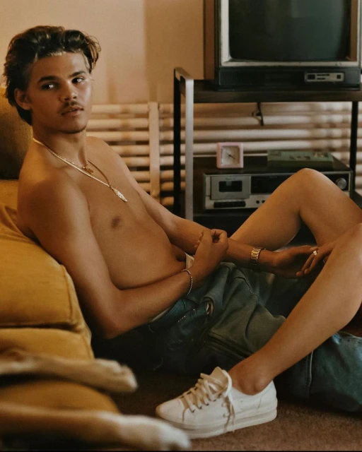 Shirtless man with jewelry sits indoors next to a vintage television and stereo. White sneakers are visible on the carpeted floor.