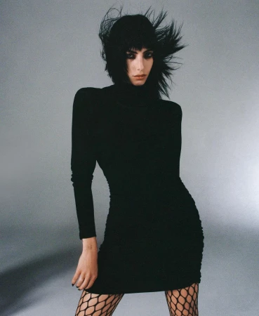 Woman in black long sleeve dress and fishnet tights with dark windswept hair against a plain grey backdrop