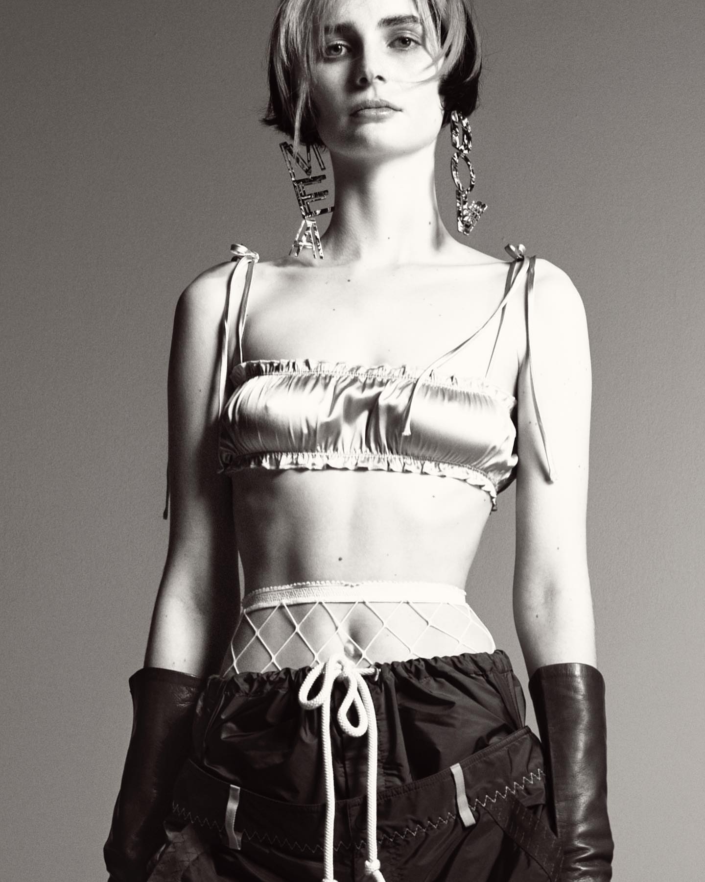 Monochrome portrait of a woman with short dark hair in a shiny crop top, dark shorts, long gloves, and large earrings against a grey backdrop.