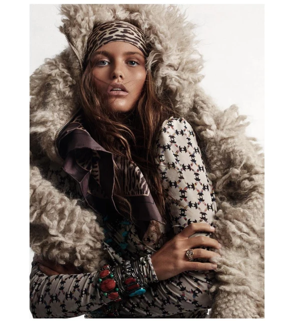 Fashion shot of a woman in a printed top and large wool coat with a leopard print headscarf, posing indoors against a white background.