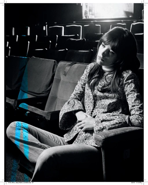 Woman with long hair in a patterned shirt sits thoughtfully in an empty theatre, a monochromatic setting with subtle blue highlights.