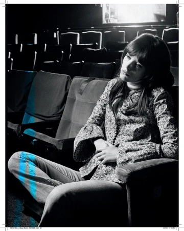 Woman with long hair in a patterned shirt sits thoughtfully in an empty theatre, a monochromatic setting with subtle blue highlights.