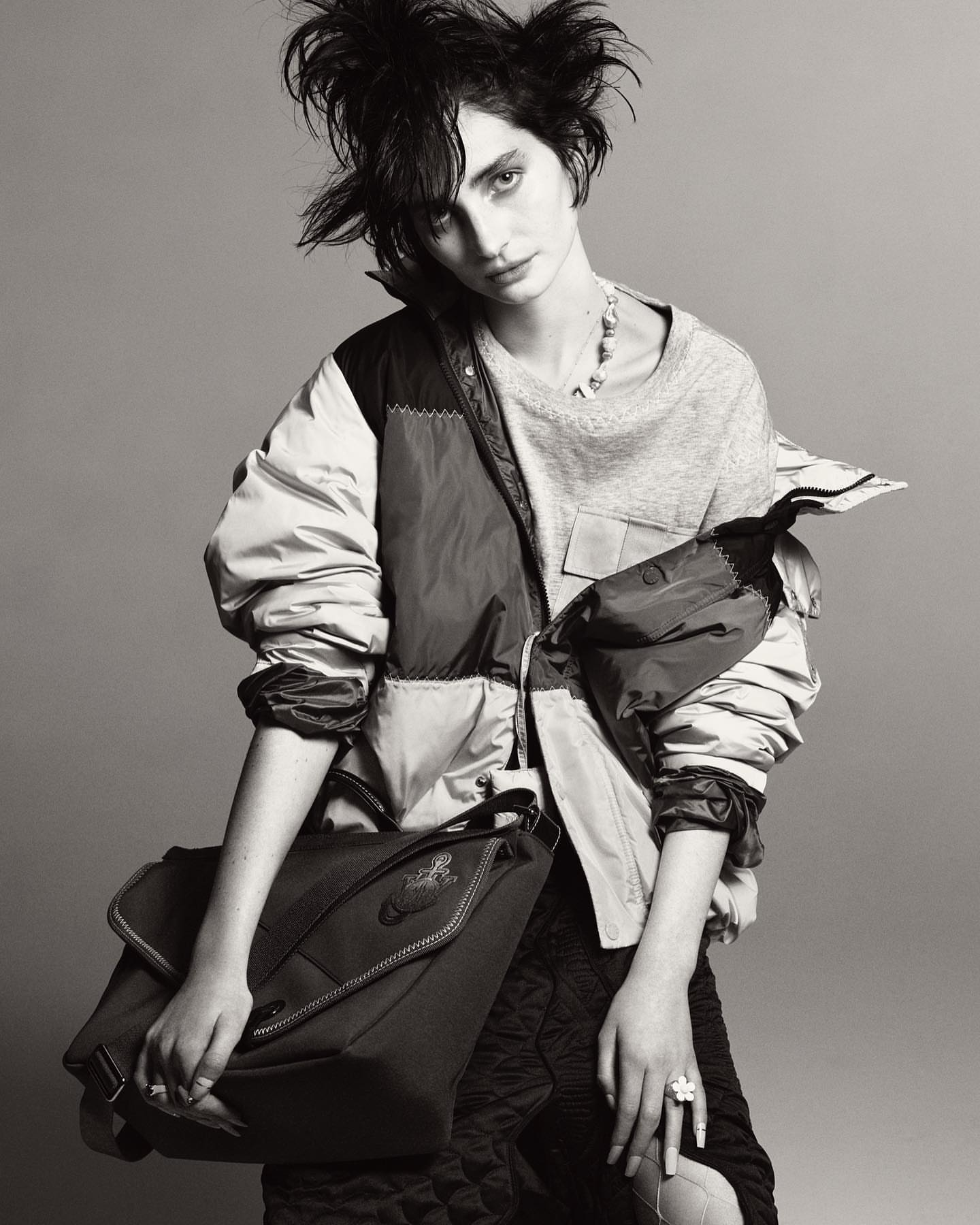 Black and white studio portrait of a woman with messy hair wearing a jacket and carrying a bag, styled in an editorial fashion.