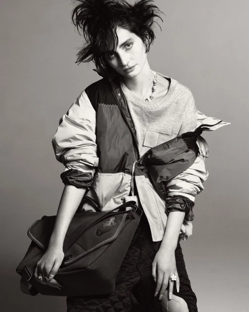 Black and white studio portrait of a woman with messy hair wearing a jacket and carrying a bag, styled in an editorial fashion.