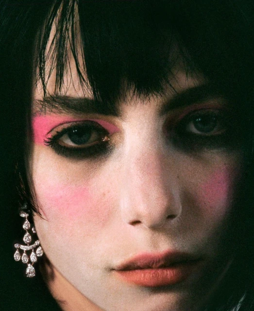 Close-up portrait of a woman with dark hair, fair skin, pink makeup on her eyelids and cheeks, and teardrop earrings.