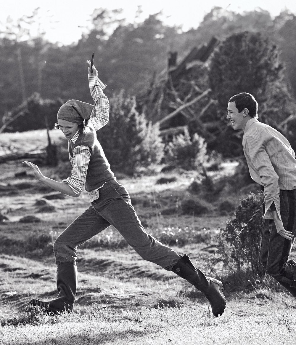 Black and white photo of a young woman joyfully leaping across a grassy field, a young man smiling behind, set against a backdrop of trees.