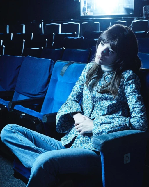 Woman with long dark hair wearing a floral top and jeans sitting in a blue theater seat looking melancholy with empty seats in the background.