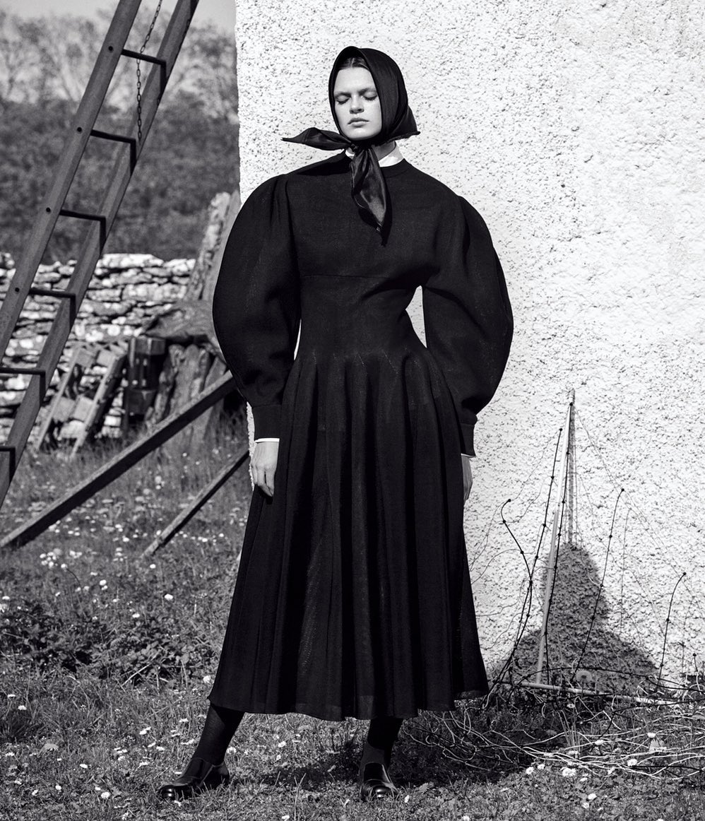 A solitary woman in a black dress and headscarf strikes a pose in a rural outdoor setting, with an old ladder and white wall in the background.