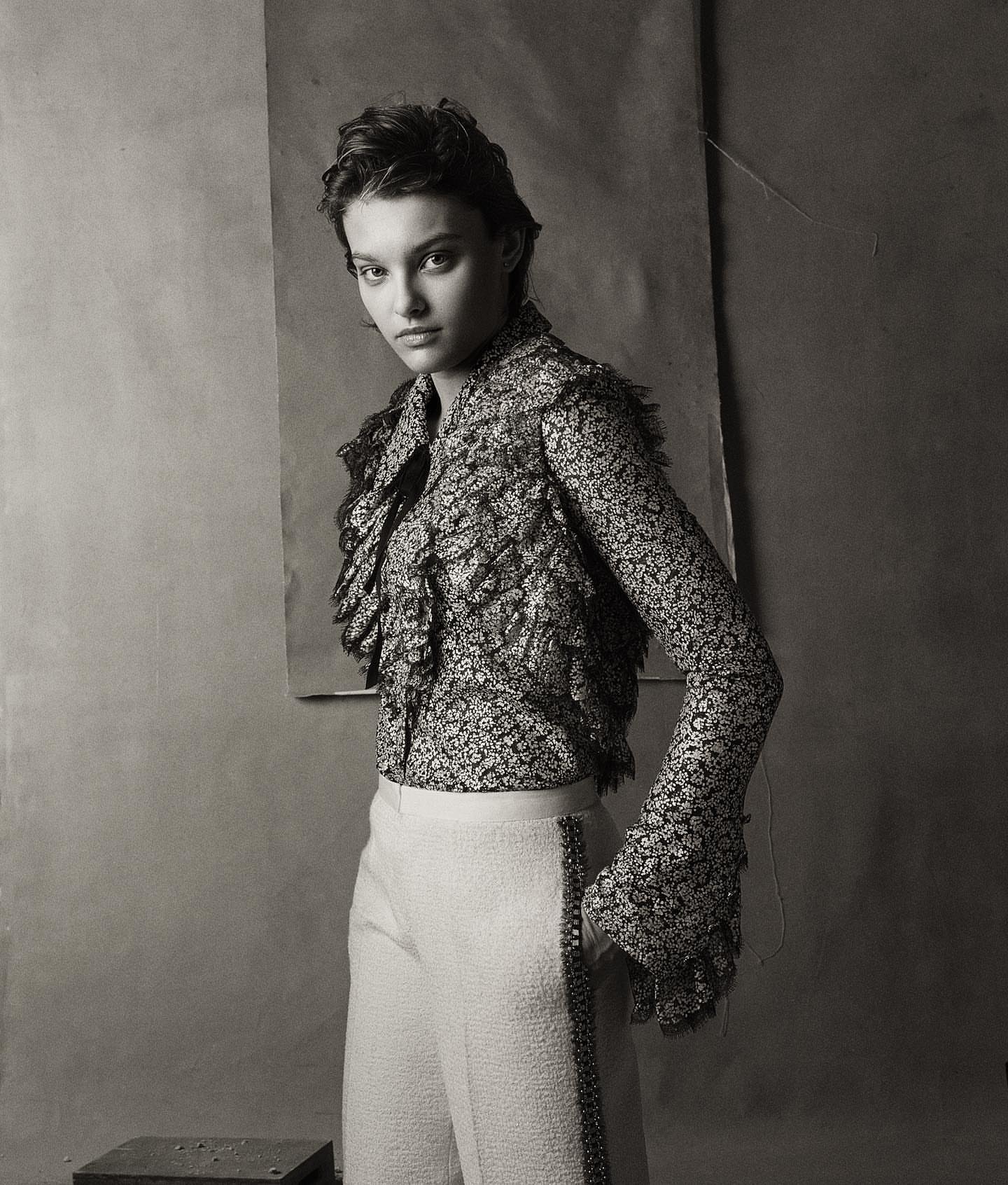 Black-and-white portrait of a young adult woman in a floral blouse and white pants, posing against a neutral backdrop.