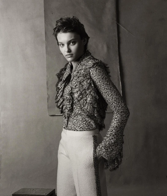 Black-and-white portrait of a young adult woman in a floral blouse and white pants, posing against a neutral backdrop.