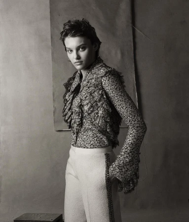 Black-and-white portrait of a young adult woman in a floral blouse and white pants, posing against a neutral backdrop.