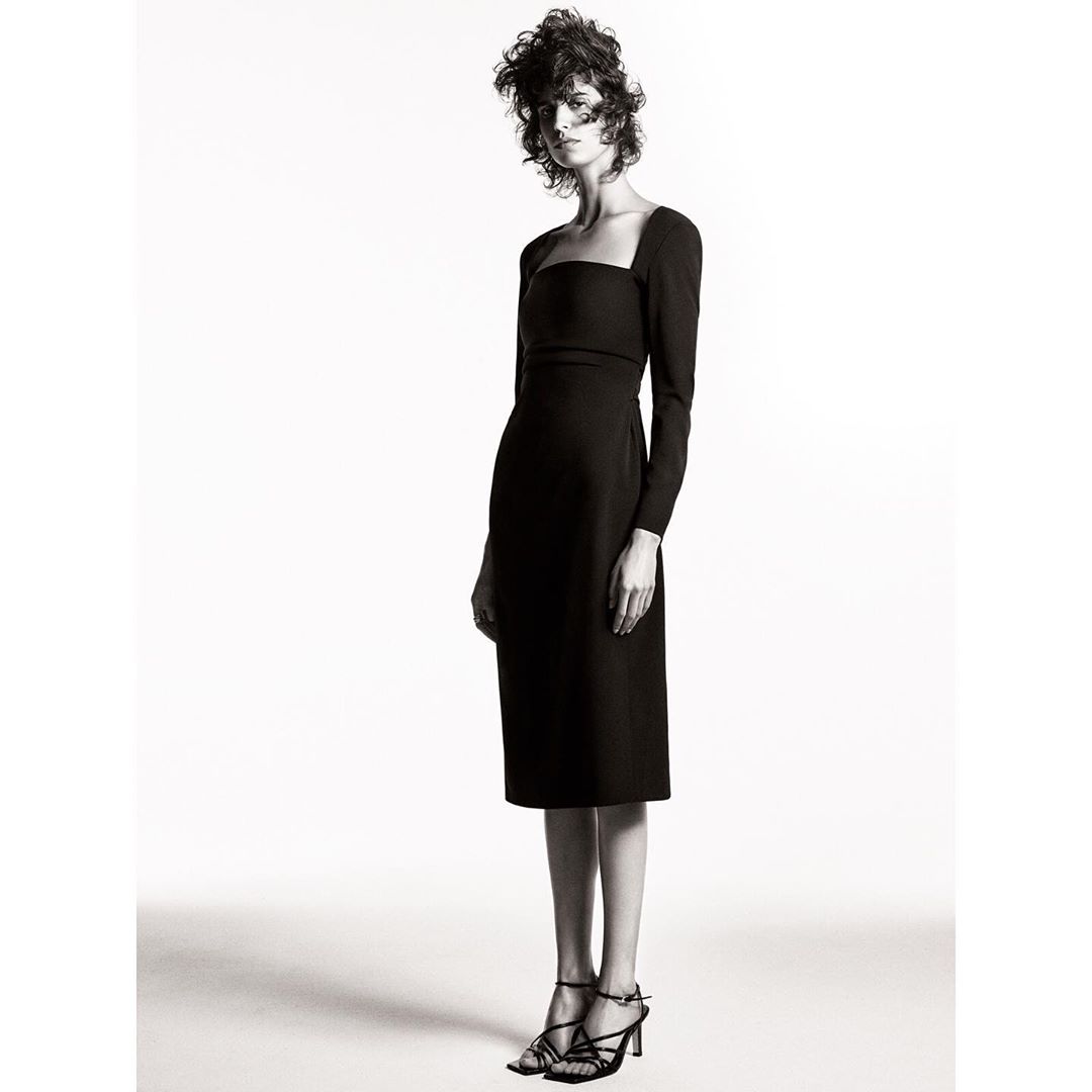 Fashion model wearing a black, long-sleeved midi dress and strappy heels in a studio shot on a white backdrop, fashion editorial.