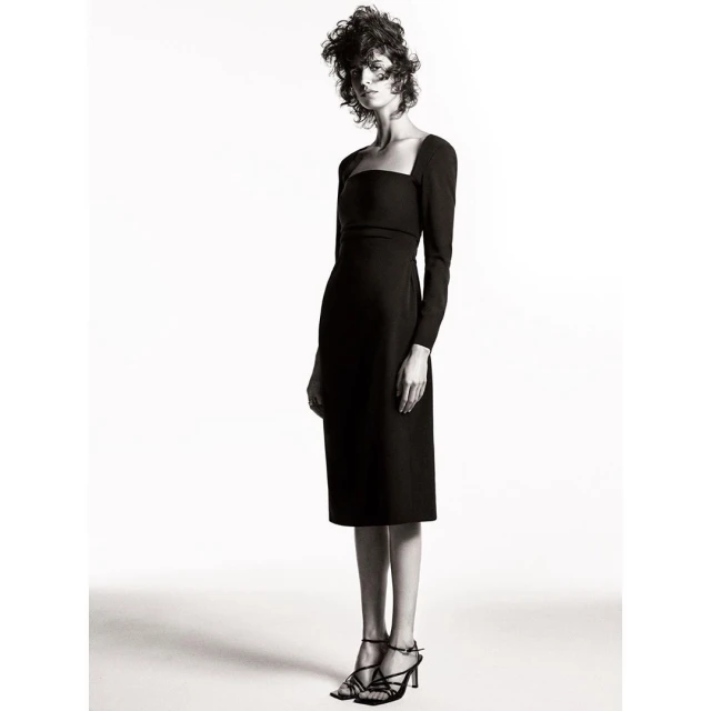 Fashion model wearing a black, long-sleeved midi dress and strappy heels in a studio shot on a white backdrop, fashion editorial.