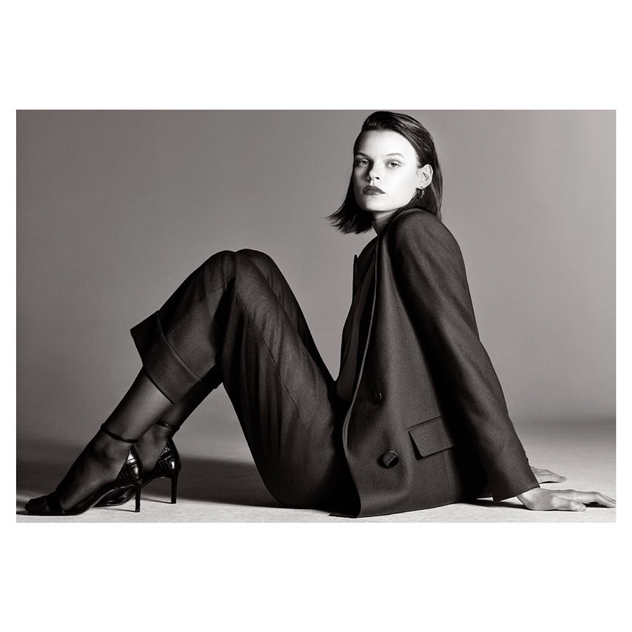 Woman poses in a grey studio wearing a black blazer, black trousers, black sheer hose, and black patent leather high heels.