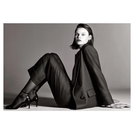 Woman poses in a grey studio wearing a black blazer, black trousers, black sheer hose, and black patent leather high heels.