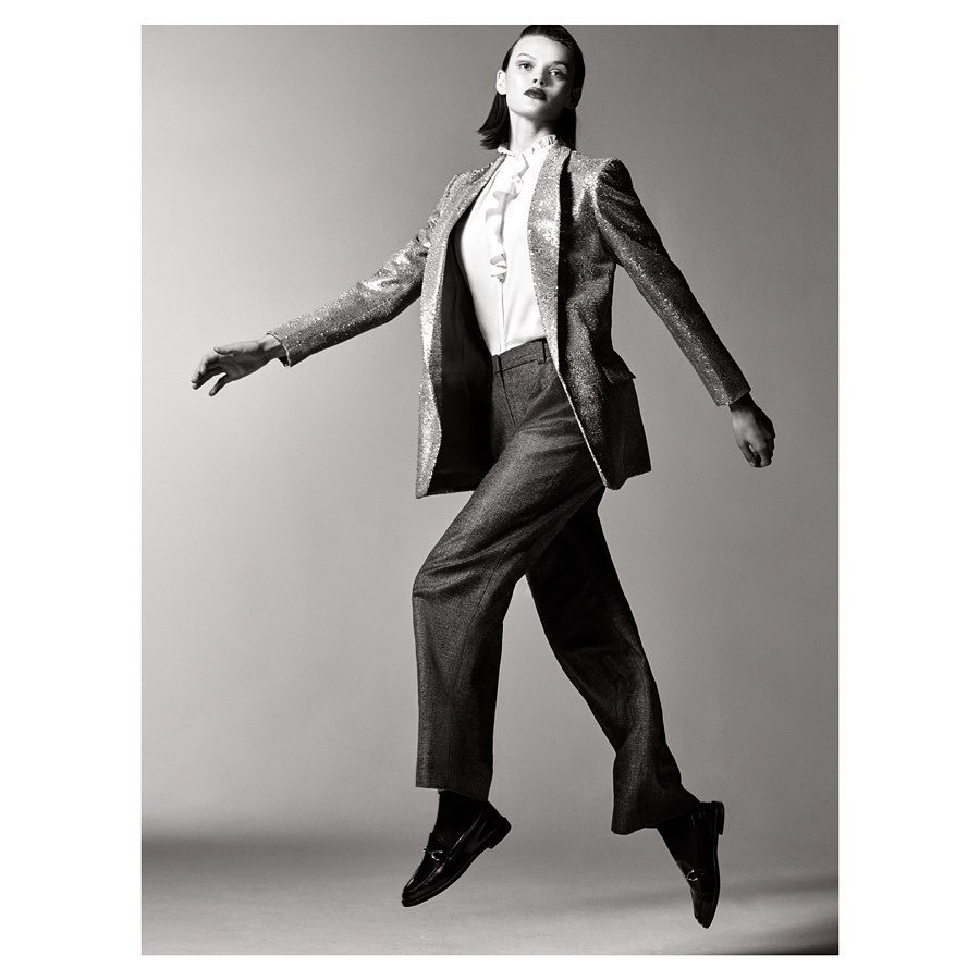 Black and white studio shot of a female fashion model in a sparkly blazer and trousers jumping in mid-air against a grey background.