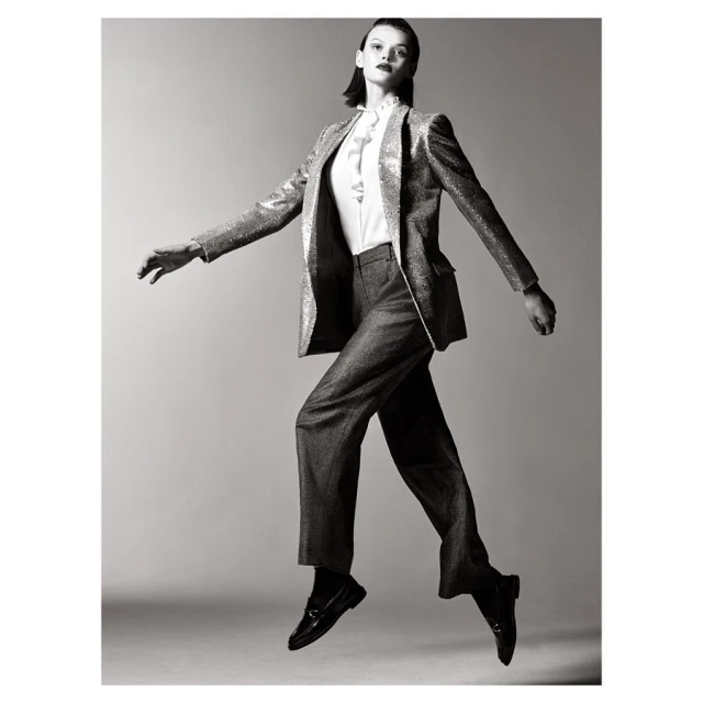 Black and white studio shot of a female fashion model in a sparkly blazer and trousers jumping in mid-air against a grey background.