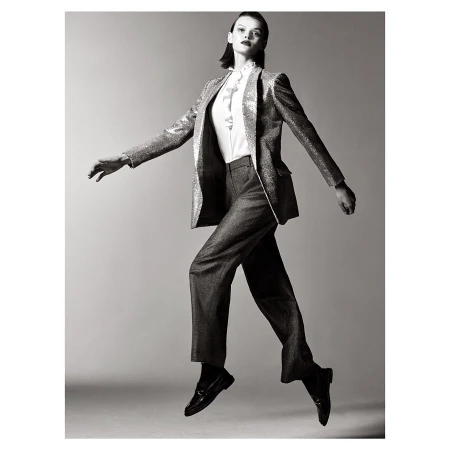 Black and white studio shot of a female fashion model in a sparkly blazer and trousers jumping in mid-air against a grey background.