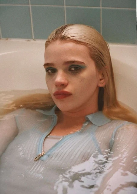 Woman with blonde hair submerged in bathtub water, wearing a light blue shirt, and dramatic blue eye makeup, looking at the camera.