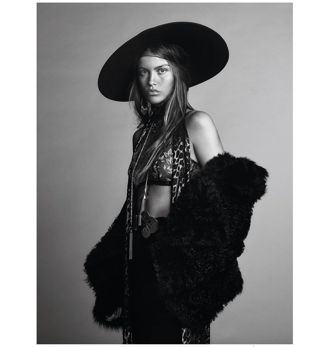 A woman with long hair poses in a studio wearing a large hat, printed top, and fur coat in a black-and-white fashion photograph.