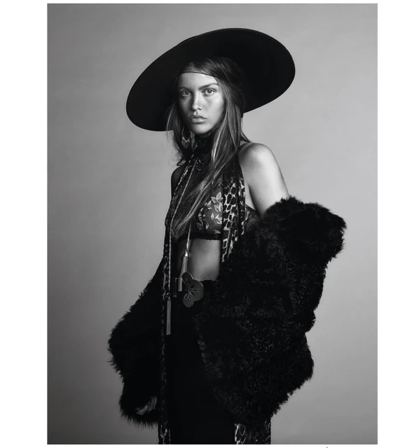 A woman with long hair poses in a studio wearing a large hat, printed top, and fur coat in a black-and-white fashion photograph.
