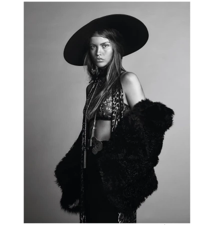 A woman with long hair poses in a studio wearing a large hat, printed top, and fur coat in a black-and-white fashion photograph.