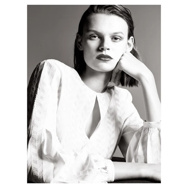Monochrome portrait of a woman with dark lipstick wearing a white blouse, resting her head on her hand in a minimalist studio.