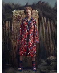 Model standing outdoors wearing a red floral coat and striped socks with an old floral mattress propped behind her.