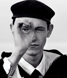 Close-up black and white portrait of a man wearing a dark beret and making a circle gesture with his hand over one eye.