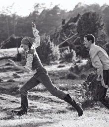 Black and white editorial image of a woman running outdoors with a man following, wearing a headscarf and tall boots.