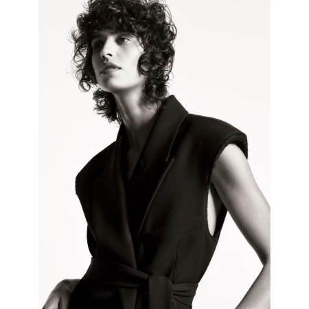 Model with curly hair wearing a dark sleeveless blazer against a plain white background by Christian Macdonald