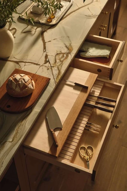 Warm-toned kitchen counter with artisanal bread, marble countertop, a drawer revealing an organized knife collection, and greenery.