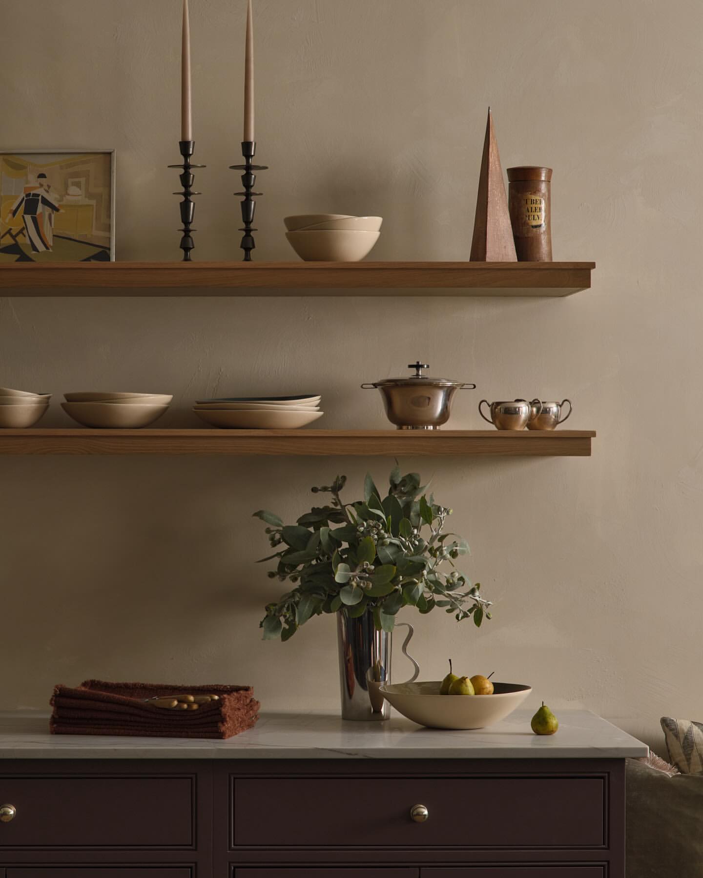 Interior still life with candles, bowls, pears, and tableware arranged on shelves and a kitchen cabinet against a beige wall.