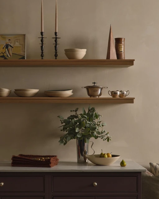 Interior still life with candles, bowls, pears, and tableware arranged on shelves and a kitchen cabinet against a beige wall.