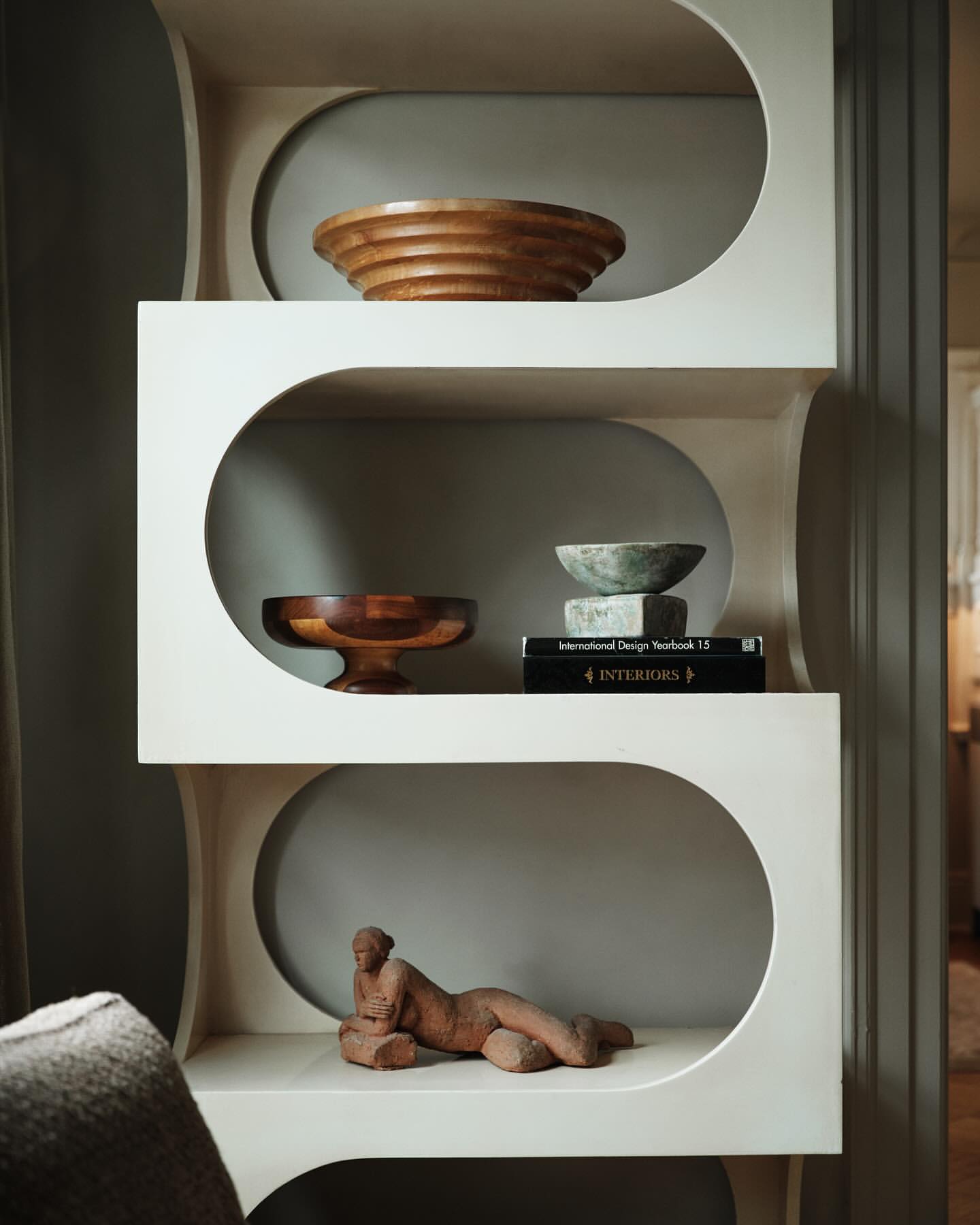 A minimalist, retro-style shelving unit displaying wooden bowls, stacked books, and a reclining figure sculpture in a neutral interior.