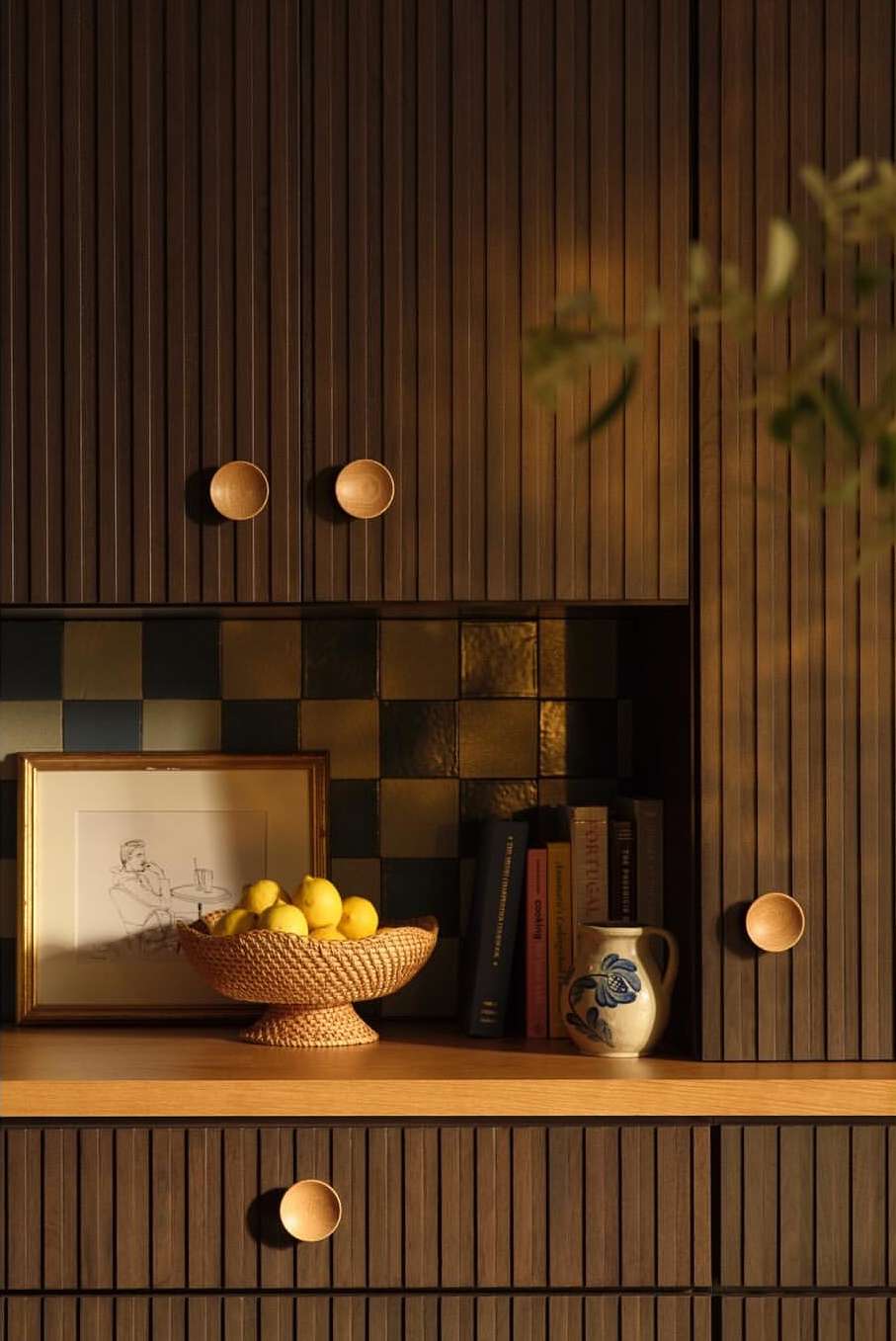 Still life featuring a woven basket with lemons, a framed picture, books, and a ceramic pitcher, set against a backdrop of dark wood cabinets.
