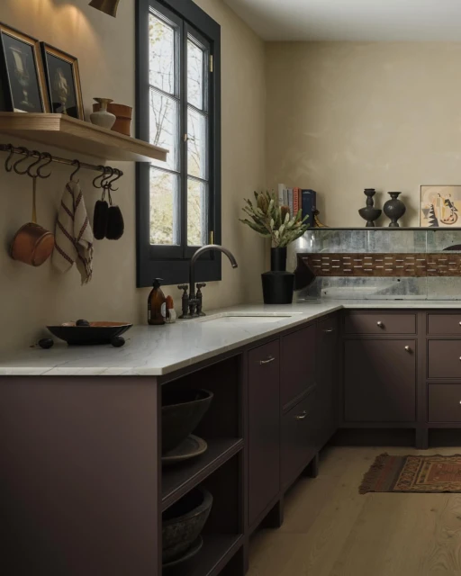 Modern kitchen design featuring deep plum cabinetry, marble countertops, copper accents, and natural lighting from a window.